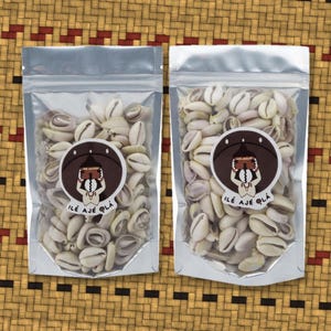 May include: Two silver-colored, resealable pouches filled with small, off-white cowrie shells. Each pouch features a circular sticker with a brown background, a stylized graphic of a figure, and the text ILÉ AJÉ OLÁ printed in black lettering.