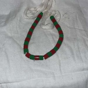 May include: A necklace featuring cylindrical beads in alternating red and green colors, strung on a white cord. The beads are arranged in a repeating pattern along the length of the cord, which is gathered in a loose loop at the top of the piece.