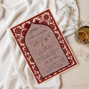 May include: A rectangular invitation card with a burgundy and cream color scheme. The card features floral and geometric patterns, with text that reads "Dalia Zayyed & Zain Khalidi" and event details. The card is on a white fabric surface.