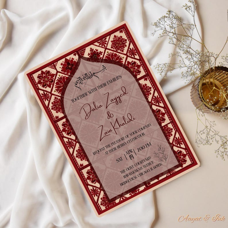 May include: A rectangular invitation card with a burgundy and cream color scheme. The card features floral and geometric patterns, with text that reads "Dalia Zayyed & Zain Khalidi" and event details. The card is on a white fabric surface.
