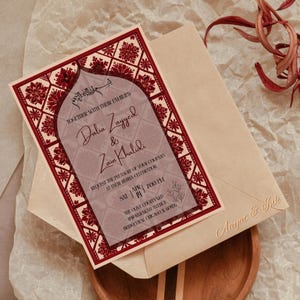 May include: A wedding invitation with a burgundy and cream color scheme. The invitation features the names Dalia Zayyed & Zain Khalidi, with the date and time of the event. The invitation is placed on a cream-colored envelope.