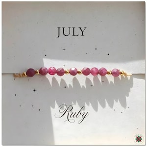 May include: A delicate bracelet featuring faceted ruby beads and small gold-colored beads. The word "July" is printed at the top, and the word "Ruby" is printed at the bottom. The bracelet is displayed on a white background.