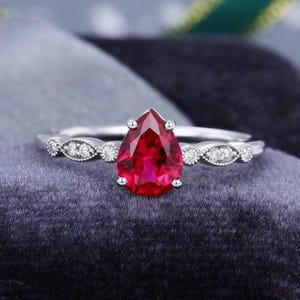 Può includere: Un anello color argento con una gemma rosso intenso a forma di pera al centro. La fascia è decorata con piccole pietre rotonde e trasparenti incastonate in una montatura metallica a forma di marquise su entrambi i lati della pietra centrale.