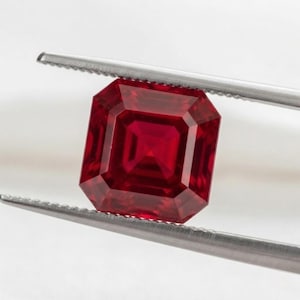 May include: A vibrant, deep red gemstone with a square step-cut facet pattern, held securely by a pair of silver-toned metal tweezers. The stone exhibits a clear, transparent quality with sharp, geometric edges and a symmetrical, multi-layered design.