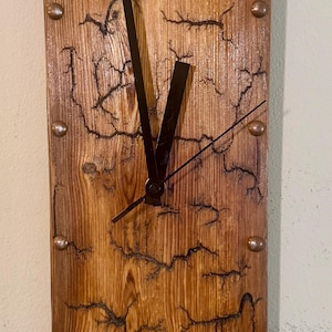 May include: A rectangular wooden clock with a natural wood grain finish. The clock face features black clock hands and copper-colored studs. The wood has a unique fractal pattern burned into it.