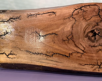May include: A rectangular wooden serving board with a natural edge. The board is a warm brown color with a glossy finish. It features intricate black fractal patterns, resembling lightning strikes, across its surface. A knot is visible in the wood grain.