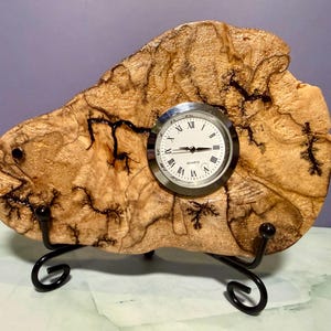 May include: A unique wooden clock with a quartz movement. The clock face is round with Roman numerals and black hands. The wooden base has a natural, light brown color with dark brown veining and is supported by a black metal stand.
