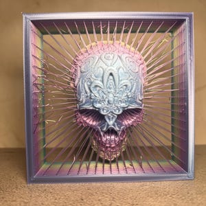 May include: A 3D printed square frame in a gradient of purple, blue, and green, containing a central skull sculpture with intricate embossed floral patterns. Thin, radiating filaments connect the skull to the inner edges of the frame.