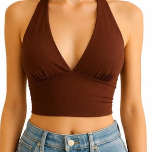 May include: A brown halter top with a deep V-neckline. The top is a solid color and appears to be made of a soft, stretchy material. The halter straps tie behind the neck. The model is wearing blue jeans.
