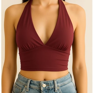 Pode incluir: Top halter cor bordô com decote em V profundo. O top é combinado com jeans azul claro. O top halter é de cor sólida e parece ser feito de um tecido liso.