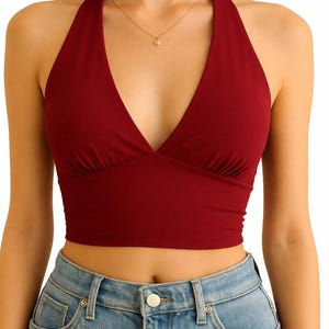 May include: A burgundy halter top with a deep V-neckline. The top is cropped and paired with light blue denim jeans. A delicate gold necklace with a small pendant is also visible.