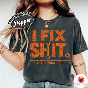 Puede incluir: Camiseta gris oscuro con el texto naranja "I FIX SHIT. THAT'S WHAT I DO" y un gráfico de llave inglesa. La camiseta tiene un aspecto vintage y desgastado, y el texto es en negrita. Marca Comfort Colors.