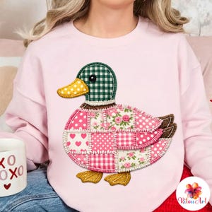 Peut inclure: Un sweat-shirt rose clair avec un motif de canard en patchwork. Le canard a une tête à carreaux verts et blancs, un bec jaune et un corps composé de patchs roses et blancs à motifs. Les pieds du canard sont dorés.