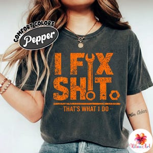 Puede incluir: Camiseta gris oscuro con el texto naranja "I FIX SHIT". Un gráfico de llave inglesa está centrado entre las palabras. Debajo está la frase "THAT'S WHAT I DO". La camiseta tiene un aspecto vintage y desgastado.