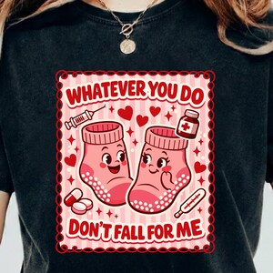 Puede incluir: Camiseta negra con un diseño gráfico. El diseño presenta dos calcetines rosas con caras, ilustraciones médicas y el texto "WHATEVER YOU DO DON'T FALL FOR ME". El diseño incluye corazones, pastillas y un termómetro.