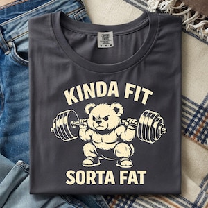 May include: Dark gray t-shirt with the text "KINDA FIT SORTA FAT" and a cartoon bear lifting weights. The shirt is folded on top of blue jeans and a plaid blanket.