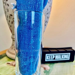May include: A clear glass tumbler with the text "Johnnie Walker" and a blue cloth inside. The cloth has a fringed edge. A black rectangular object with the words "Keep Walking" is visible in the background.