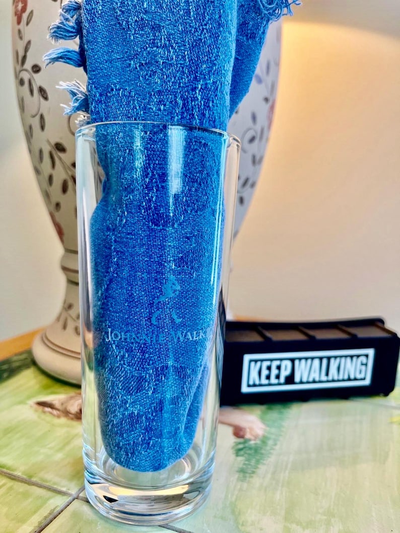 May include: A clear glass tumbler with the text "Johnnie Walker" and a blue cloth inside. The cloth has a fringed edge. A black rectangular object with the words "Keep Walking" is visible in the background.