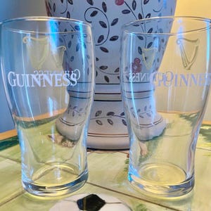 May include: Two clear pint glasses with the Guinness logo and harp emblem are displayed. The glasses are empty and sit in front of a decorative vase with floral patterns. The glasses are on a table with a soccer ball.