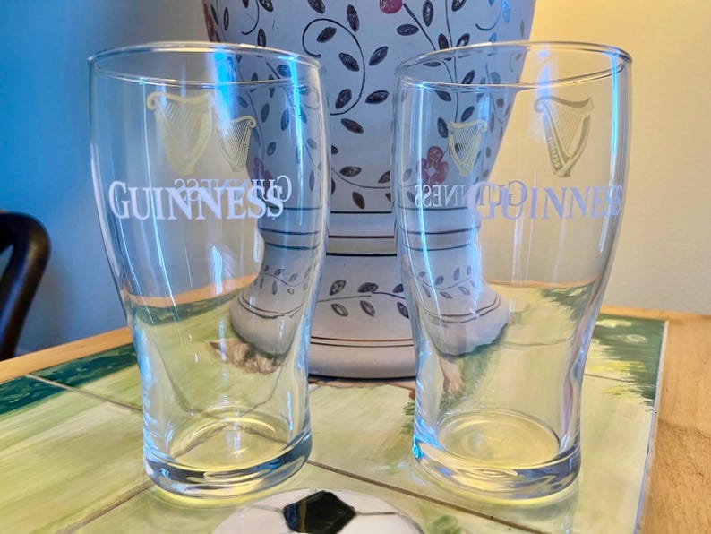 May include: Two clear pint glasses with the Guinness logo and harp emblem are displayed. The glasses are empty and sit in front of a decorative vase with floral patterns. The glasses are on a table with a soccer ball.