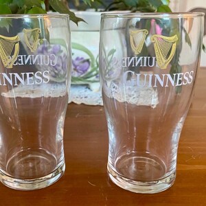 May include: Two clear Guinness pint glasses. Each glass has the Guinness logo in gold and white. The logo includes a harp and the word "Guinness". The glasses are empty and sit on a wooden surface.