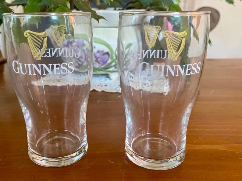 May include: Two clear Guinness pint glasses. Each glass has the Guinness logo in gold and white. The logo includes a harp and the word "Guinness". The glasses are empty and sit on a wooden surface.