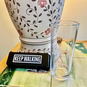 May include: A clear, tapered glass and a black rectangular object with the words "KEEP WALKING" in white text. A white vase with a floral design is in the background.