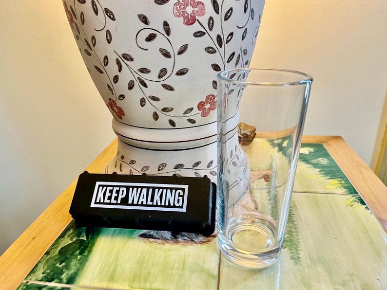 May include: A clear, tapered glass and a black rectangular object with the words "KEEP WALKING" in white text. A white vase with a floral design is in the background.