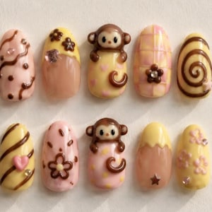 Op de afbeelding: Een set van tien kunstnagels met diverse ontwerpen. De nagels hebben een mix van roze, gele en bruine kleuren, met decoratieve elementen zoals krullen, bloemen, harten en een apenfiguur. Sommige nagels hebben een glanzende afwerking.