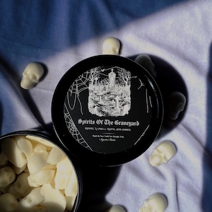 May include: A black candle tin with the text "Spirits Of The Graveyard" and a graveyard illustration. The tin is surrounded by small, white skull-shaped wax melts. The candle's scent is coffee, vanilla, clove, and amber.