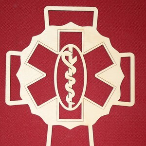 May include: A wooden Star of Life symbol, a medical emblem, cut from light-colored wood. The design features a six-pointed star with a staff of Asclepius in the center, set against a red background.