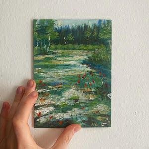 May include: An original landscape painting depicting a forest scene with a stream and red flowers. The artwork is painted with green, blue, and white brushstrokes. The painting is held by a hand against a white wall.