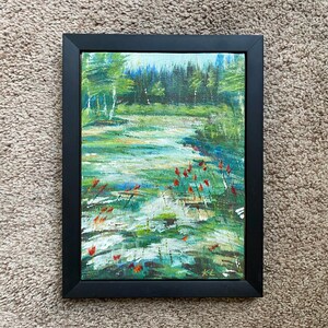 May include: A framed landscape painting depicts a serene waterscape with a forest backdrop. The artwork features a palette of greens, blues, and touches of red, with visible brushstrokes. The black frame adds a contrasting border to the nature-themed art.