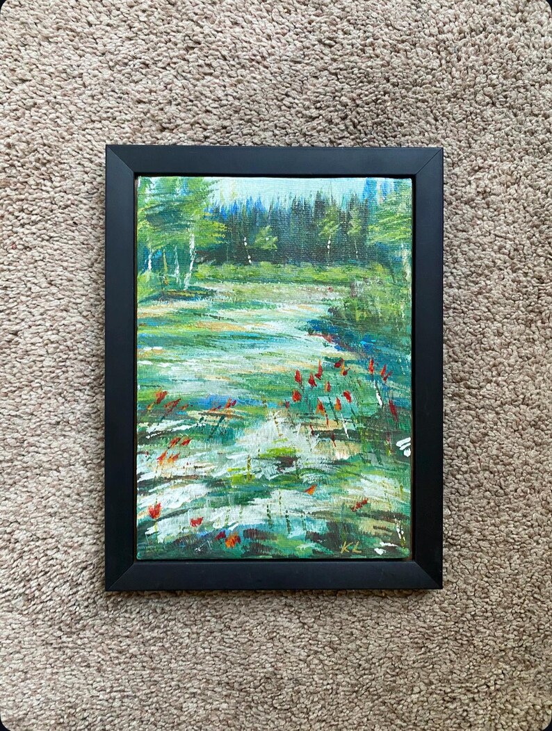 May include: A framed landscape painting depicts a serene waterscape with a forest backdrop. The artwork features a palette of greens, blues, and touches of red, with visible brushstrokes. The black frame adds a contrasting border to the nature-themed art.
