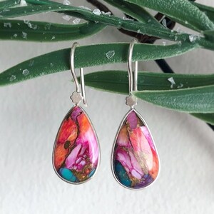 Op de afbeelding: Een paar druppelvormige oorbellen met een zilverkleurig montuur. De oorbellen hebben een kleurrijke steen met roze, oranje, turquoise en gouden tinten. Ze hangen voor een groene, bladerrijke achtergrond.