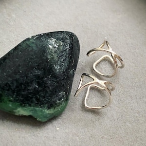 May include: Two silver ear cuffs with an abstract design, beside a dark green and black stone. The ear cuffs have a curved, open design. The stone has a rough texture and a green edge.