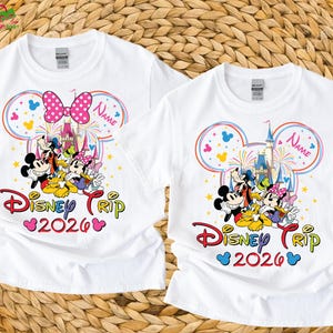 May include: Two white t-shirts with a colorful Disney Trip 2026 design. The design features Mickey Mouse, Minnie Mouse, Donald Duck, Goofy, and Daisy Duck in front of a castle. One shirt has a pink bow, the other has a name field.