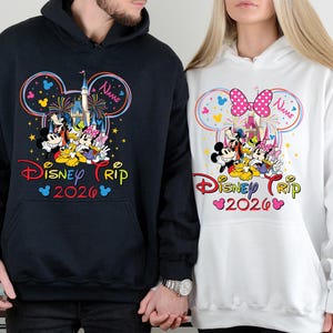 Op de afbeelding: Twee hoodies, een zwarte en een witte, met een kleurrijk Disney Trip 2026-ontwerp. Het ontwerp bevat Mickey en Minnie Mouse, een kasteel, vuurwerk en de tekst "Disney Trip 2026".