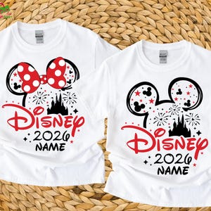 May include: Two white t-shirts with a Disney-themed design. One shirt features a red polka-dot bow, while both display black Mickey Mouse ears, a red "Disney" logo, the year "2026", and space for a name, with a castle and fireworks.