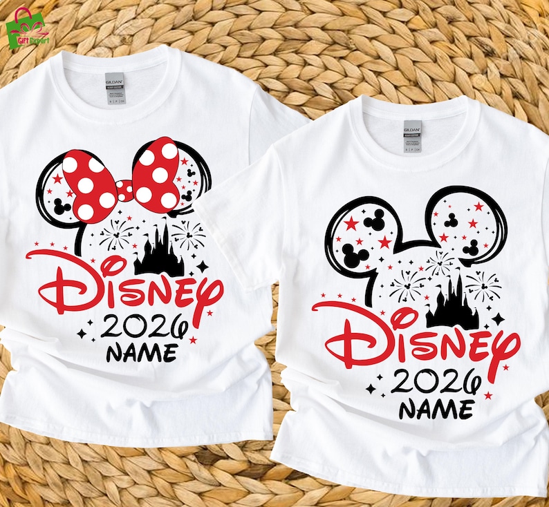May include: Two white t-shirts with a Disney-themed design. One shirt features a red polka-dot bow, while both display black Mickey Mouse ears, a red "Disney" logo, the year "2026", and space for a name, with a castle and fireworks.