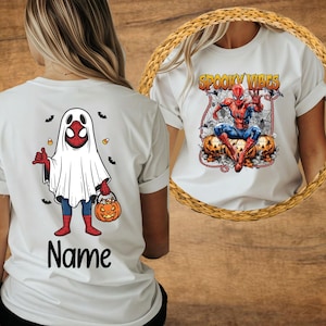 May include: White t-shirts with Halloween-themed graphics. One shirt features a ghost costume with a Spider-Man mask, holding a pumpkin bucket, with the word "Name" below. The other shirt displays Spider-Man with the text "Spooky Vibes" and pumpkins.