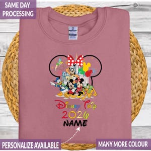 Pode incluir: Camisola rosa empoeirada com um gr&aacute;fico colorido de personagens da Disney. O design inclui Mickey e Minnie Mouse, Pato Donald, Pateta e Pluto. O texto diz "Disney Trip 2020" e "NAME". A camisola &eacute; personaliz&aacute;vel e est&aacute; dispon&iacute;vel em muitas cores.