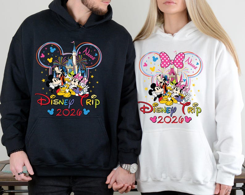 Op de afbeelding: Twee hoodies, een zwarte en een witte, met een kleurrijk Disney Trip 2026-ontwerp. Het ontwerp bevat Mickey en Minnie Mouse, een kasteel, vuurwerk en de tekst "Disney Trip 2026".