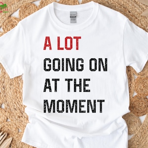 May include: White t-shirt with the phrase "A LOT GOING ON AT THE MOMENT" printed in red and black block letters. The shirt is made of a soft material and has a crew neck.
