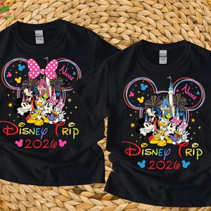 May include: Black t-shirts featuring a colorful Disney Trip 2026 design. The design includes Mickey Mouse, Donald Duck, and other characters, with a castle and fireworks. One shirt has a pink bow, the other has a name.