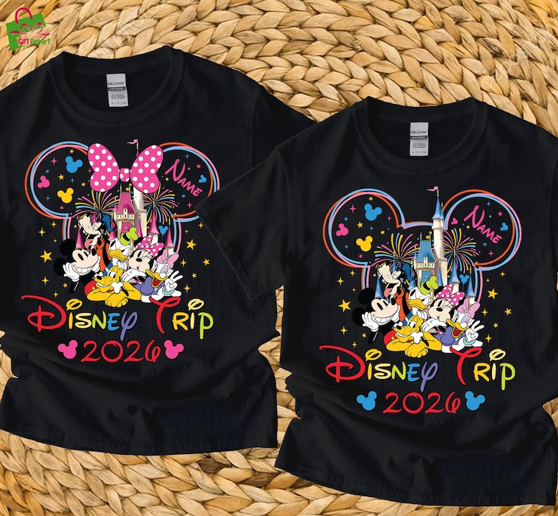 May include: Black t-shirts featuring a colorful Disney Trip 2026 design. The design includes Mickey Mouse, Donald Duck, and other characters, with a castle and fireworks. One shirt has a pink bow, the other has a name.