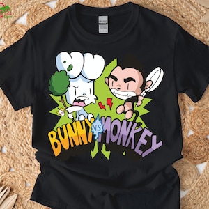 Könnte beinhalten: Schwarzes T-Shirt mit einem Cartoon-Motiv eines weißen Hasen, der einen Ast hält, der einem braunen Affen mit einem Löffel gegenübersteht. Der Text "BUNNY vs MONKEY" ist in bunten Buchstaben dargestellt.