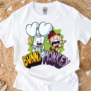 May include: White t-shirt with a cartoon graphic of a bunny and a monkey. The bunny is white with large, heart-shaped ears, holding a sword and shield. The monkey is brown and red, holding a green object. The text "BUNNY vs MONKEY" is below the characters.