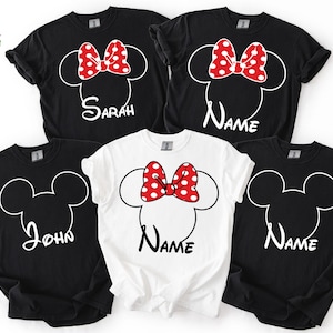 May include: Five black and white t-shirts with a Minnie Mouse design. The design includes a red bow with white polka dots and a name in white script. One white shirt has the same design. The shirts are casual wear.