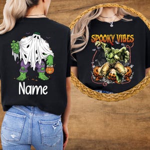 May include: Black t-shirts with Halloween-themed graphics. One shirt features a green character dressed as a ghost holding a pumpkin and the word "Name". The other shirt displays a green character with the text "Spooky Vibes" and pumpkins.